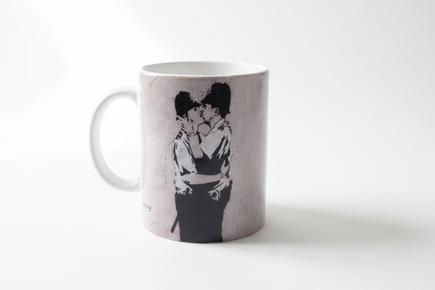 Banksy Kissing Police ceramic mug on clean white surface, bright lifestyle product photography